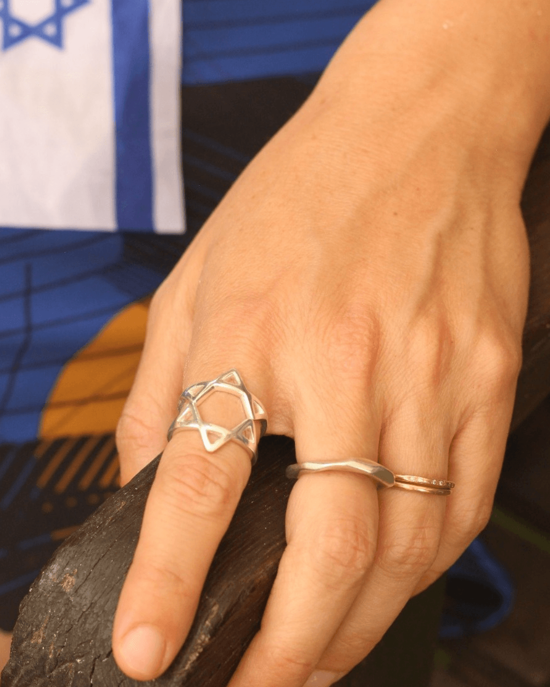 A hand resting on dark stone, wearing the sterling silver ring on one finger, with an Israeli flag in the background.